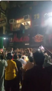 Crowd outside Udham Restaurant, with people visible at an upper-floor opening in the incident video