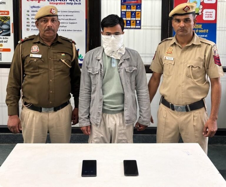 Two Delhi Police personnel beside a masked man, with two mobile phones on a table